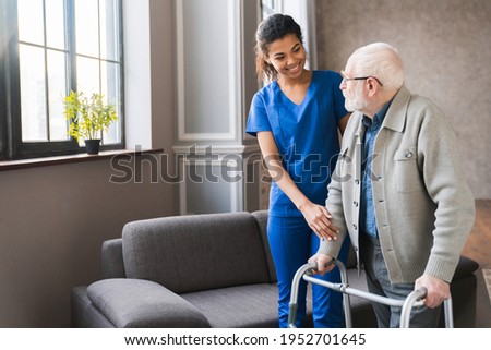 Portrait of an african young nurse helping old elderly disable man grandfather to walk using walker equipment in the bedroom. Senior patient of nursing home moving with walking frame and nurse support