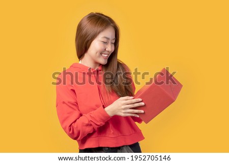 Portrait of casual young happy smiling Asian woman hold gift box. Beautiful young Asian girl holding red gift box over yellow background. Cute Asian girl with red jacket holding gift box.