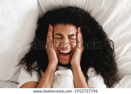 Scared stressed young african girl crying in her white bed. Top view of dark skinned woman scared of nightmare, lies in bed with white bedclothes, afraids of staying alone, slept bad at night