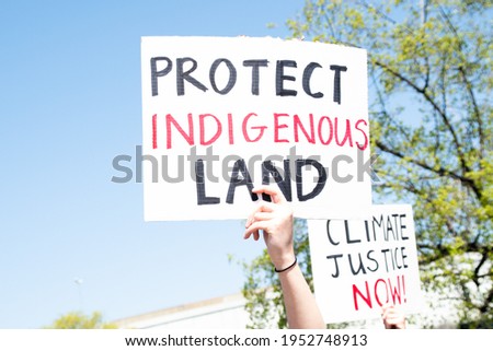Caucasian hand holding sign reading "Protect Indigenous Land" at a protest in response to fossil fuel pipelines being routed through protected Native American reservations and danger of polluted water Royalty-Free Stock Photo #1952748913