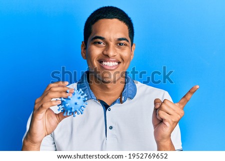 Young handsome hispanic man holding virus toy smiling happy pointing with hand and finger to the side 