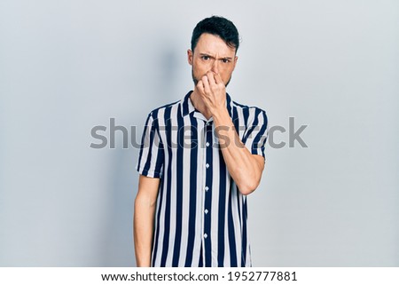 Young hispanic man wearing casual clothes smelling something stinky and disgusting, intolerable smell, holding breath with fingers on nose. bad smell 