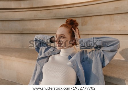 Beauty portrait of stylish girl posing against stairs background. Adorable young ginger wearing light clothes, smiling and touching head with hands outdoors