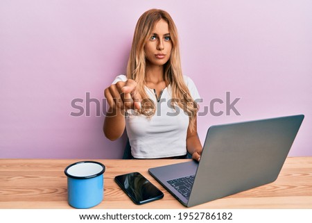 Beautiful blonde young woman working using computer laptop pointing with finger to the camera and to you, confident gesture looking serious 