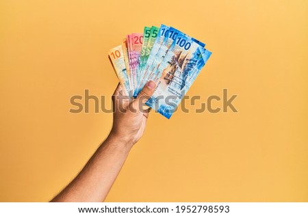 Hand of hispanic man holding swiss franc banknotes over isolated yellow background.