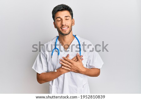 Young handsome man wearing doctor uniform and stethoscope smiling with hands on chest, eyes closed with grateful gesture on face. health concept. 