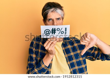Young hispanic man covering mouth with insult message paper with angry face, negative sign showing dislike with thumbs down, rejection concept  Royalty-Free Stock Photo #1952812315