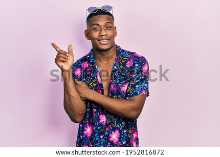 Young black man wearing hawaiian shirt and sunglasses with a big smile on face, pointing with hand and finger to the side looking at the camera. 
