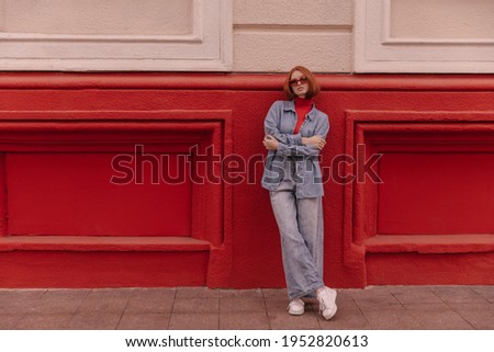 Full-length photo of young lady posing against bright background. Gorgeous red-haired woman wearing sunglasses, blue shirt, jeans and white sneakers, crossing arms outdoors