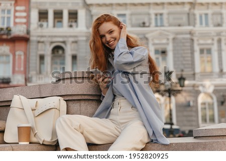 Lifestyle portrait of young long-haired foxy, sitting on stairs against buildings background. Businesswoman in stylish shirt and pants, holding phone, smiling outdoors Royalty-Free Stock Photo #1952820925
