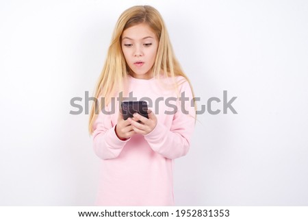 beautiful caucasian little girl wearing pink sweater over white background looks with bugged eyes, holds modern smart phone, receives unexpected message from friend, reads reminder.