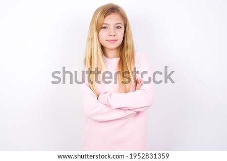 Waist up shot of  self confident beautiful caucasian little girl wearing pink sweater over white background has broad smile, crosses arms, happy to meet with colleagues.