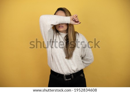 Pretty woman with long hair covering eyes with arm smiling cheerful and funny against yellow wall