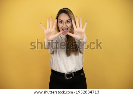 Pretty woman with long hair showing and pointing up with fingers number ten while smiling confident and happy against yellow wall