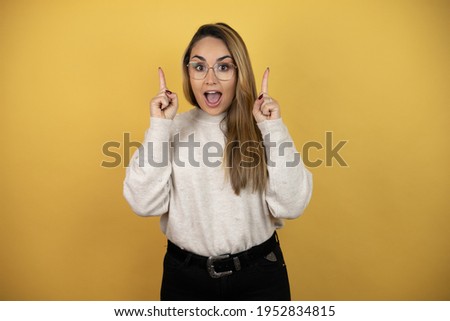 Pretty woman with long hair amazed and surprised looking at the camera and pointing up with fingers and raised arms against yellow wall