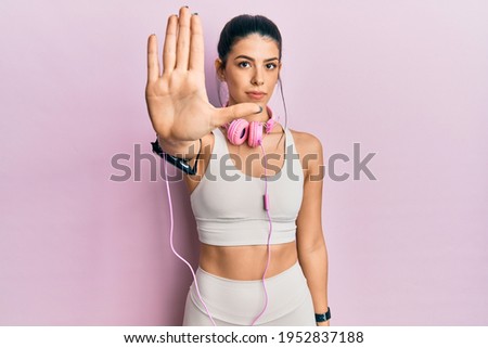 Young hispanic woman wearing gym clothes and using headphones doing stop sing with palm of the hand. warning expression with negative and serious gesture on the face. 