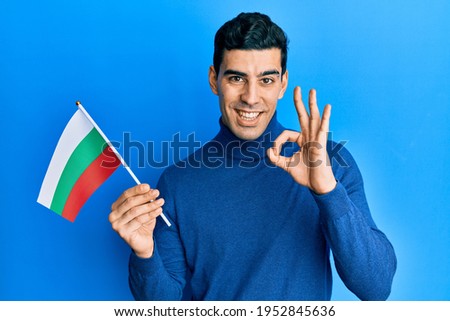 Handsome hispanic man holding bulgarian flag doing ok sign with fingers, smiling friendly gesturing excellent symbol 