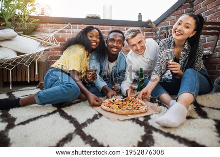 Four multicultural people in stylish clothes taking pieces of tasty pizza from box, smiling and looking at camera. Cheerful friends drinking alcoholic cocktails and eating snacks on party.