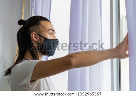 Long-haired young man with mask, looking out the window protecting himself from the new wave of covid-19.