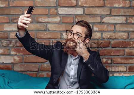 Portrait bright young gentleman with glasses, a mustache and a beard looking into a smartphone and taking a selfie against a brick wall background. Selfie concept