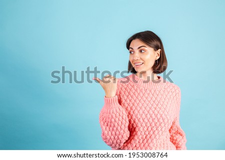 Young brunette in warm pink sweater isolated on blue background smiling point left with thumb finger 