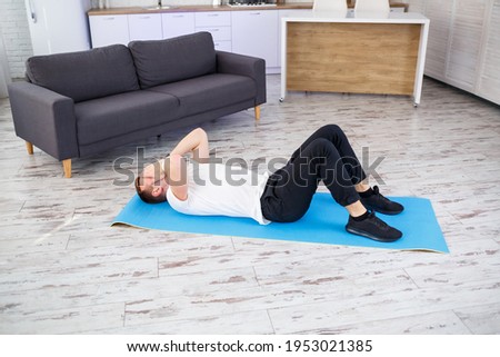 A strong athletic man in a T-shirt and doing press exercises at home in his spacious and bright apartment with a minimalistic interior. Doing sports at home.