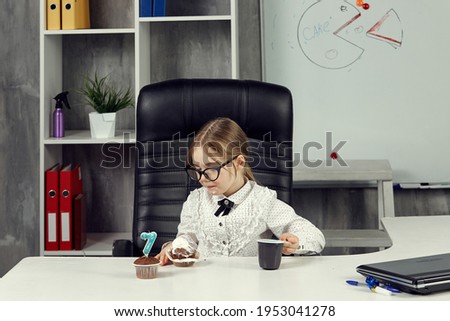 A little girl dressed as a businesswoman is sitting at an office desk eating a cupcake. The concept of business children. Children Are The Bosses. A board with a picture of a cake in the background.