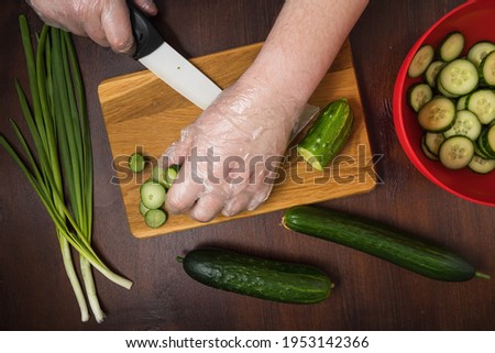 Chef is cooking vegetable salad on cutting board. Dark Photography of vegetables on brown wooden table. Flat lay of kitchen tabletop with chopping board, cucumber and spring onion. View of cook hands.