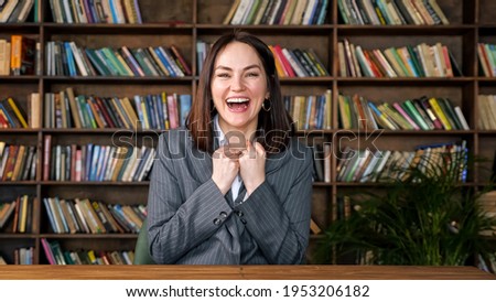 Successful businesswoman in white blouse under grey jacket raises hands with happiness sitting at table against coloured books on racks