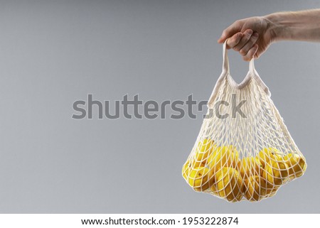 Man's hand holds string bag with lemons on solid gray background. Eco-friendly spring concept.