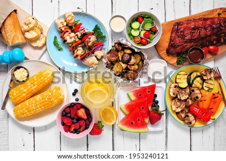 Summer BBQ or picnic food concept. Assortment of grilled meats, vegetables, fruits, salad and potatoes. Top down view table scene with a white wood background.
