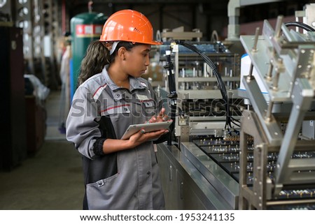 A young black manufacturing worker controls an assembly line in a factory, making notes on a tablet. Young woman in hard hat watches the work process Royalty-Free Stock Photo #1953241135