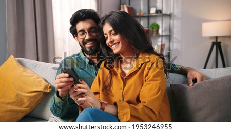Portrait of cheerful positive young lovely couple smiling spending time together at home sitting on sofa typing on smartphone, searching internet using social network app on cellphone, family concept Royalty-Free Stock Photo #1953246955