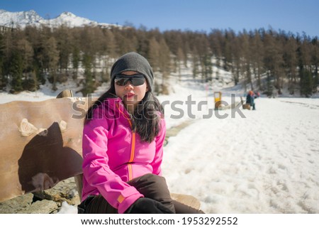 freezing Winter holidays - young happy and beautiful Asian Chinese woman on bench at frozen lake landscape surrounded by snow mountains enjoying Swiss Alps getaway