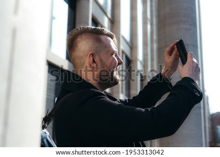 Cheerful cute guy in a black coat makes photos on the phone while walking around the city
