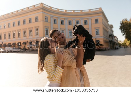 Lifestyle portrait of young couple against city landmark background at sunny warm day. Cute dark-haired girl in yellow outfit kissing and hugging boyfriend, holding little black dog