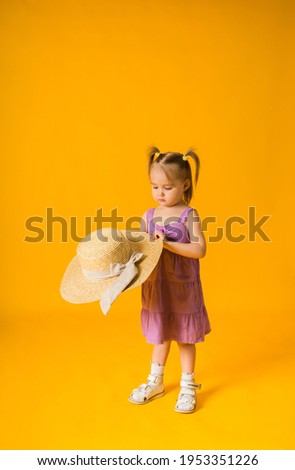 a little girl in a purple sundress holds a straw hat on a yellow background with space for text
