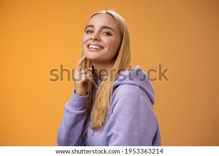 Attractive sassy self-assured caucasian blond female in purple hoodie turning camera touching chin cheeky smile camera check out interesting thing gazing forward intrigued, standing orane background