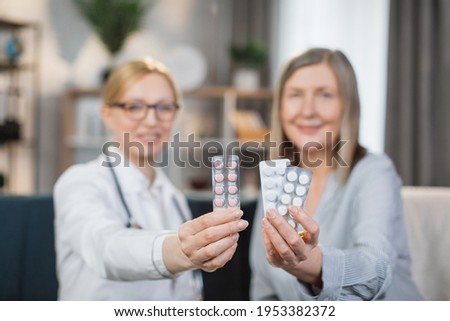 Blur view of pretty young blond woman doctor sitting on the couch at home while visiting her senior lady patient, showing blister packs with tablets pills to camera and smiling. Focus on medicines
