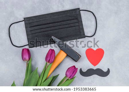 A hammer, flowers, a mask and a paper mustache with a heart lie on a gray concrete background, close-up top view.