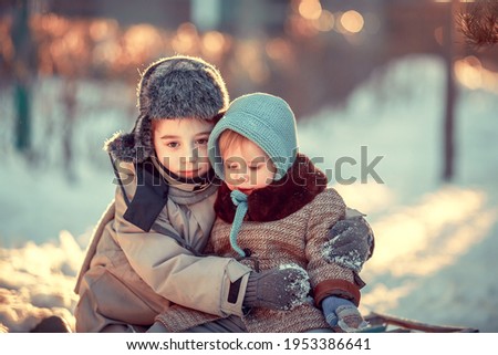 Cute little boy in earflaps hat and his baby sister in blue bonnet and retro coat are sitting on the old sledge in the winter park in Russia during the sunset. Image with selective focus and toning