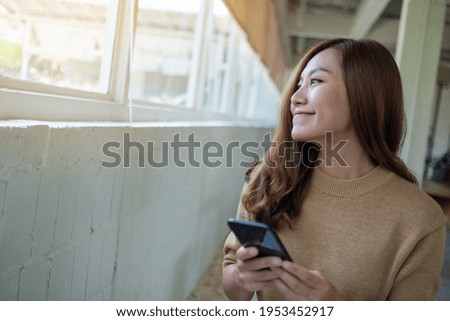Portrait image of a beautiful young asian woman looking outside the window while holding and using mobile phone 