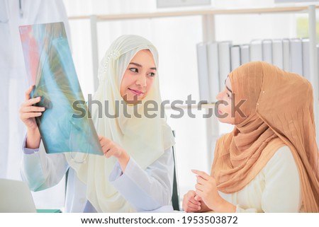 Attractive smiling young adult Muslim doctor wearing cream hijab sitting and pointing the chest x-ray film to show and explain the abnormalities of lungs to patient.