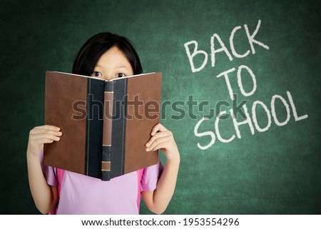 Picture of a cute schoolgirl peeping out of a book while standing in the classroom