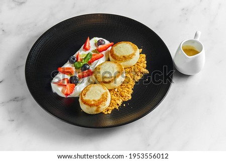 An ideal breakfast - Cottage cheese pancakes with white sauce, berries, strawberries and crunch in a black plate on a marble background. Ukrainian syrniki