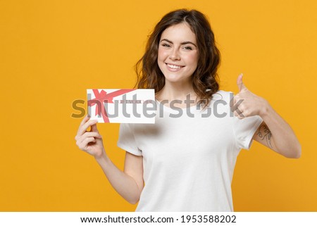 Young happy smiling fun caucasian student woman 20s wear white basic t-shirt point index finger on gift voucher flyer mock up show thumb up like gesture isolated on yellow background studio portrait