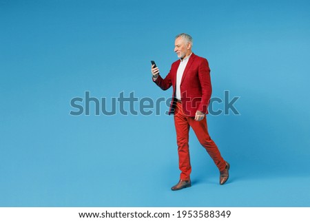 Full length side view of smiling elderly gray-haired mustache bearded business man wearing red jacket suit using mobile cell phone typing sms message isolated on blue color background studio portrait Royalty-Free Stock Photo #1953588349