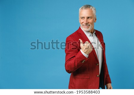 Side view of smiling elderly gray-haired mustache bearded business man wearing red jacket suit standing pointing thumb aside on mock up copy space isolated on blue color background studio portrait Royalty-Free Stock Photo #1953588403