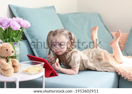 A little girl watches cartoons on a tablet computer and eats chips, fast food. Computer addiction, lifestyle