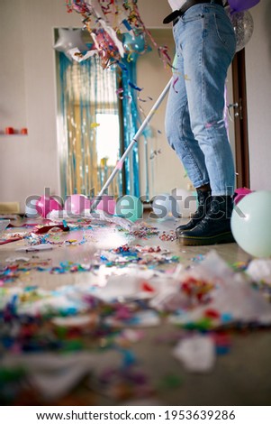 Woman with pushbroom cleaning mess of floor in room after party confetti, morning after party celebration, housework, cleaning service Royalty-Free Stock Photo #1953639286